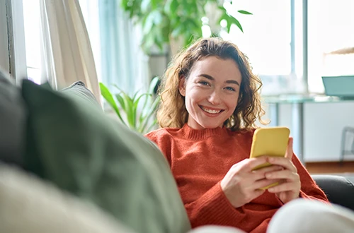 Smiling young woman relaxing on sofa and using smartphone
